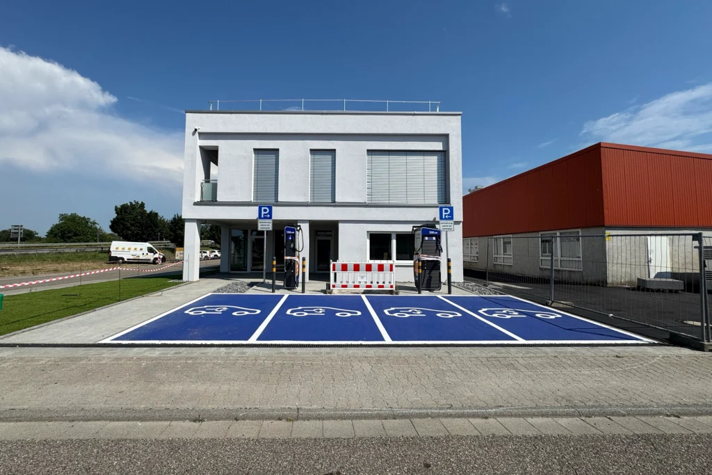 Modernes Bürogebäude mit vier blau markierten Parkplätzen für Elektroautos und zwei Ladestationen vor dem Eingang, bei sonnigem Wetter.