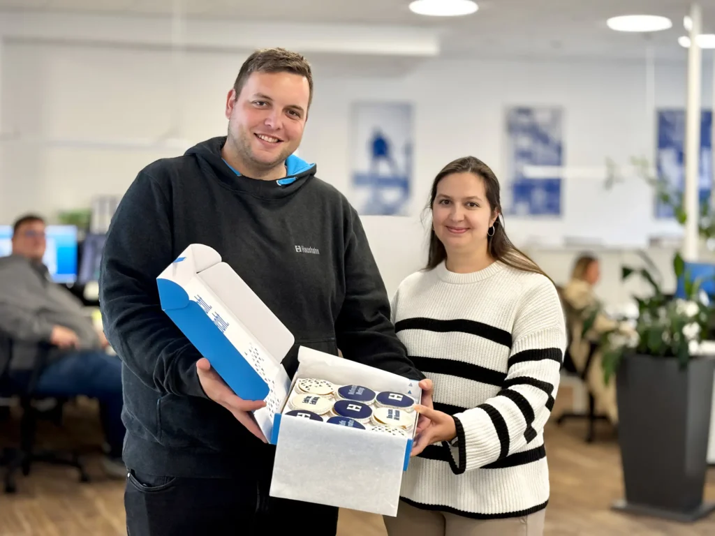 Zwei Mitarbeitende bei orbau halten im Büro eine Geschenkbox mit Muffins, die von unserem Partner Haushahn überreicht wurden.