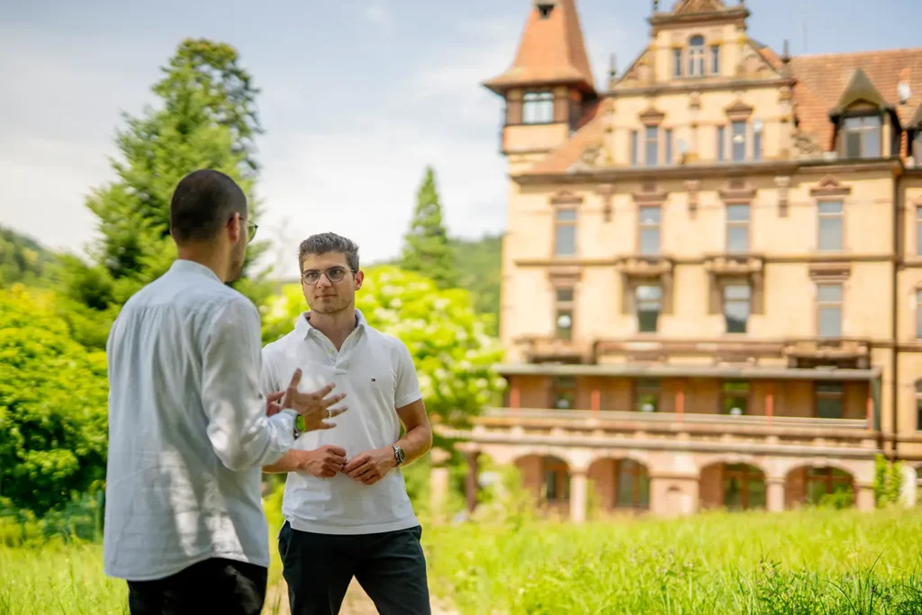 Die beiden Architekten des Schloss Rothschild stehen vor dem denkmalgeschützen Gebäude bei strahlendem Sonnenschein.