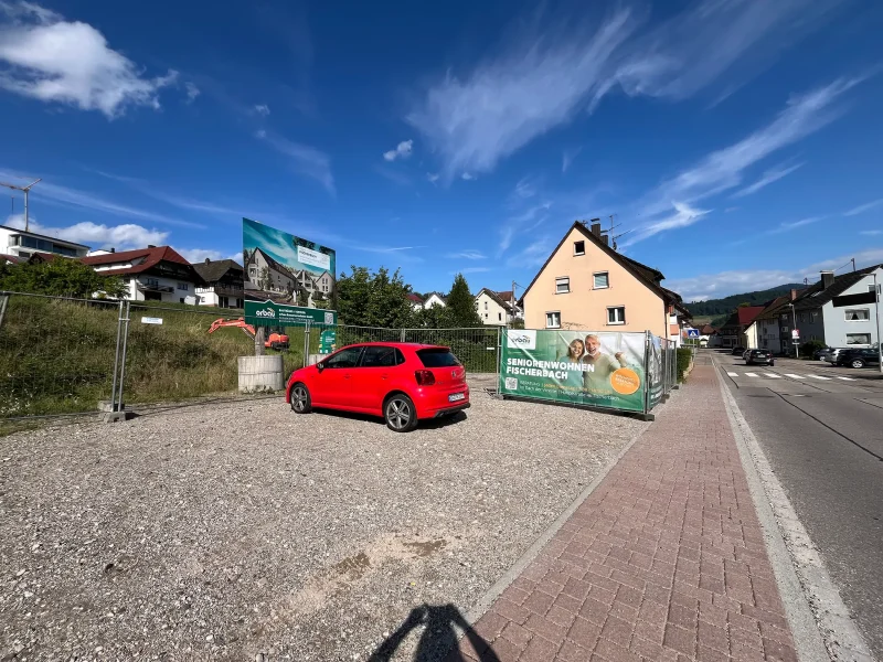 Blick auf das noch unbebaute Grundstück des Projekts „Seniorenwohnen Fischerbach“ mit Werbebanner, Wiese und angrenzenden Häusern