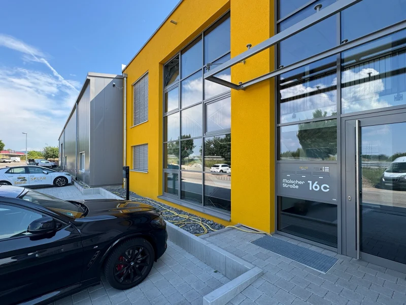 Gelbes Bürogebäude mit Glasfassade und danebenliegender grauer Halle in Durmersheim, mit parkenden Autos.